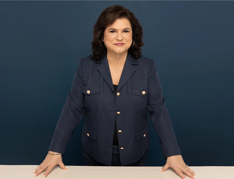 Misti Zaruches Barnett standing at a white table, smiling, wearing a dark blue jacket with silver buttons.